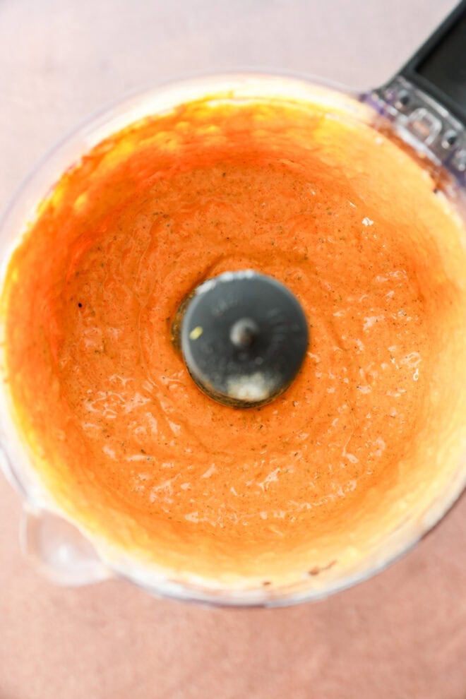 A food processor bowl containing smooth, orange-colored sauce or dip, viewed from above. The sides of the bowl are coated with the mixture, and the black blade is visible in the center.