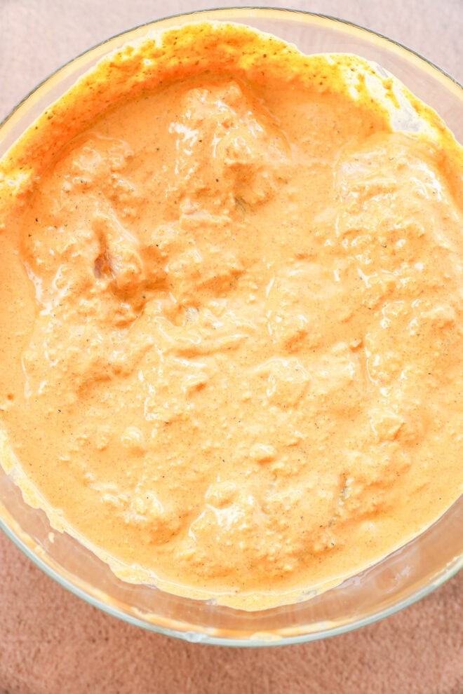 A close-up of a glass bowl filled with a thick, creamy orange-colored marinade with visible spices and small chunks, set on a light textured surface.