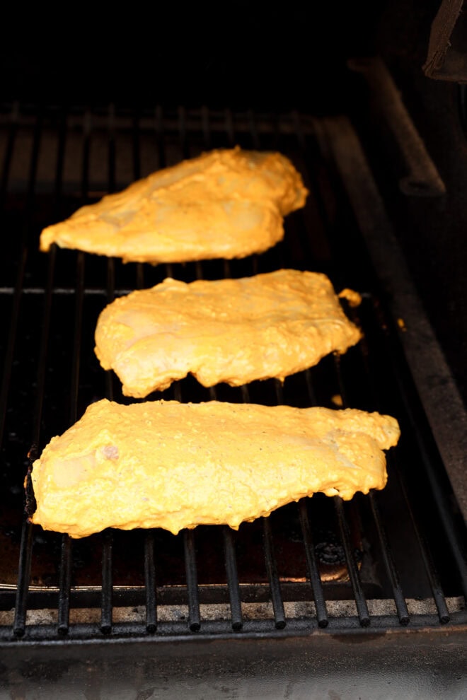 Three pieces of chicken coated in a yellow marinade are cooking on a grill, with grill marks visible on the grates beneath them.