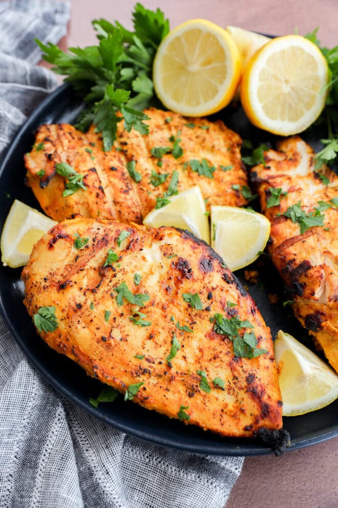 Grilled, seasoned chicken breasts garnished with fresh chopped herbs, served on a black plate with lemon wedges and parsley, next to a gray and white striped cloth.