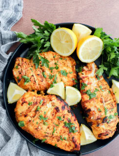 A black plate with three grilled chicken breasts garnished with chopped parsley and surrounded by fresh parsley sprigs and lemon wedges, set on a textured surface with a striped cloth.