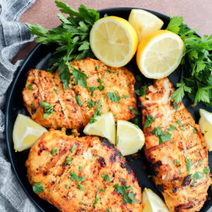 A black plate with three grilled chicken breasts garnished with chopped parsley and surrounded by fresh parsley sprigs and lemon wedges, set on a textured surface with a striped cloth.