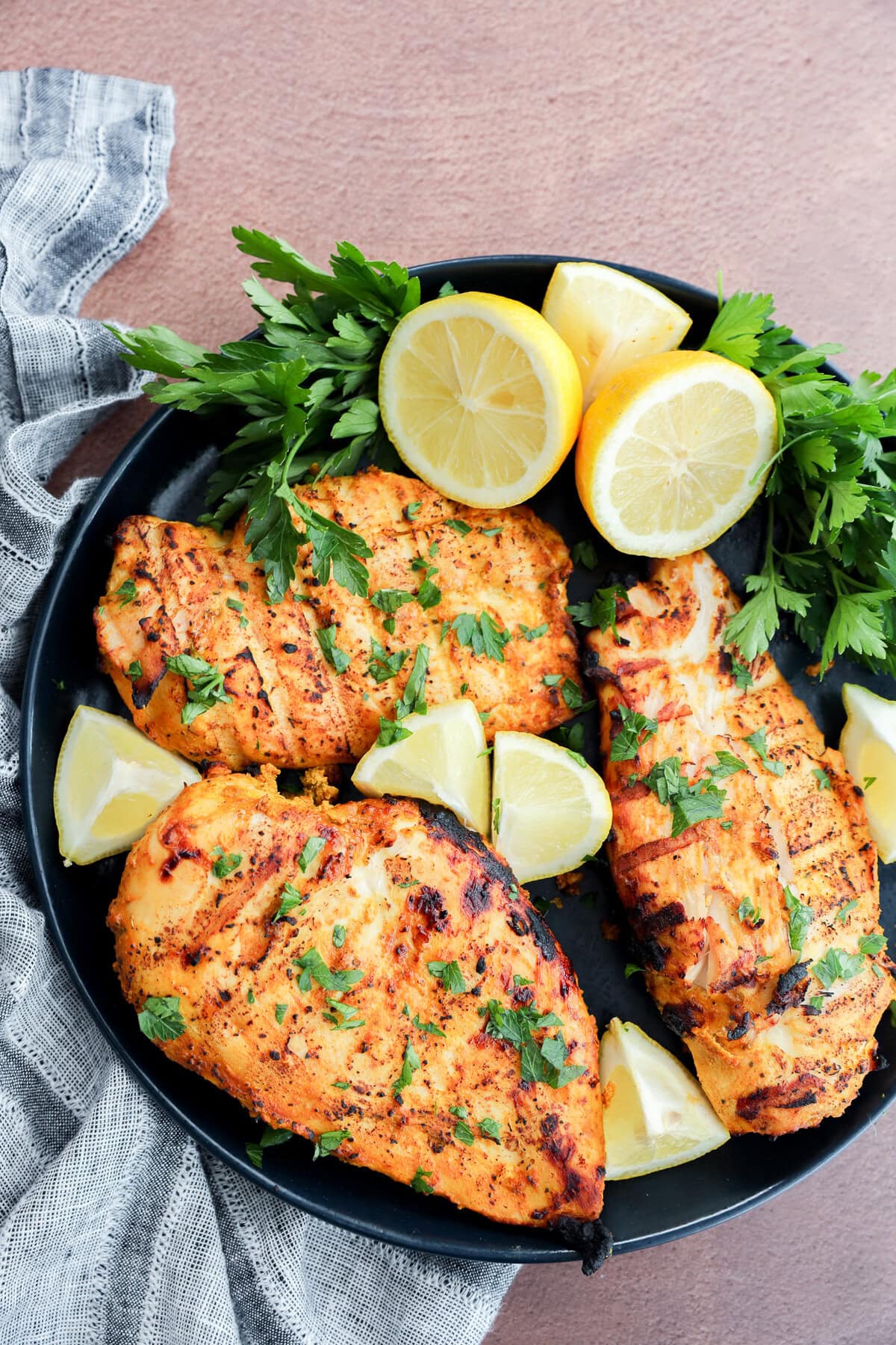 A black plate with three grilled chicken breasts garnished with chopped parsley and surrounded by fresh parsley sprigs and lemon wedges, set on a textured surface with a striped cloth.