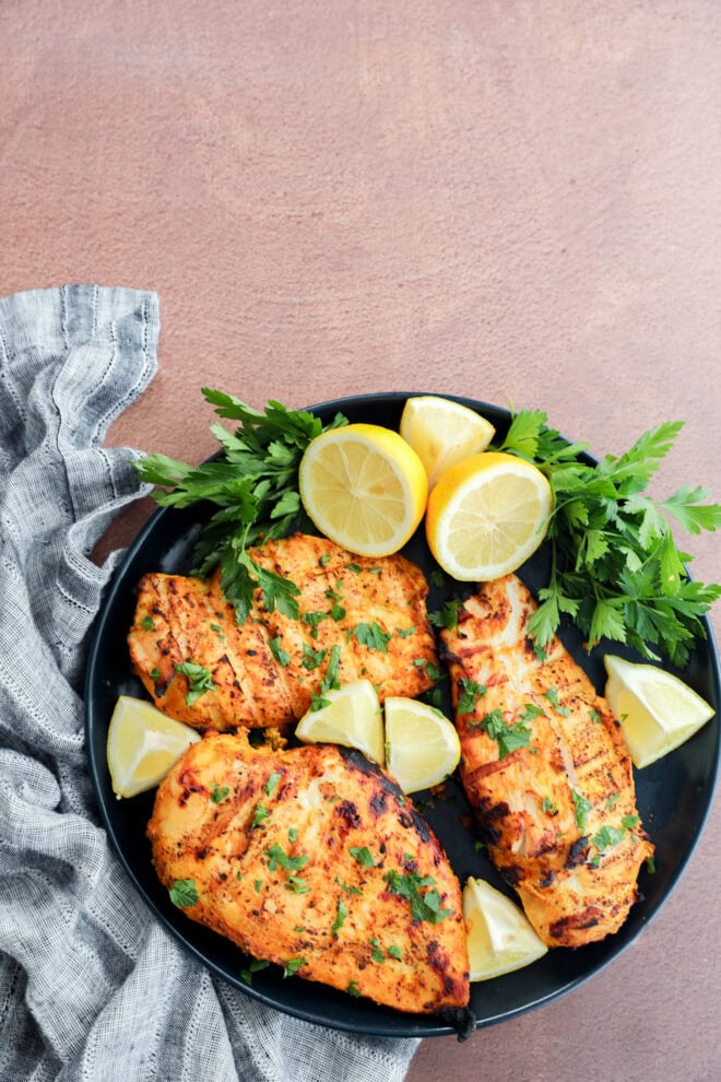 A black plate with three grilled chicken breasts garnished with chopped herbs, surrounded by lemon wedges and fresh parsley, placed on a textured brown surface next to a gray cloth napkin.