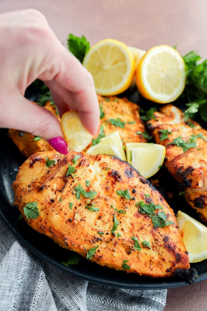 A hand squeezes a lemon wedge over grilled chicken breasts garnished with chopped herbs, with more lemon wedges and fresh parsley in the background on a black plate.