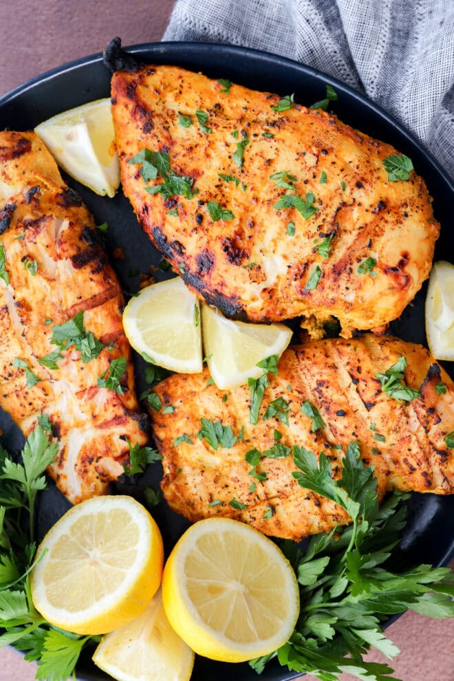 Three grilled, seasoned chicken breasts garnished with fresh parsley are arranged on a black plate with lemon wedges and parsley sprigs. A gray napkin is partially visible in the background.