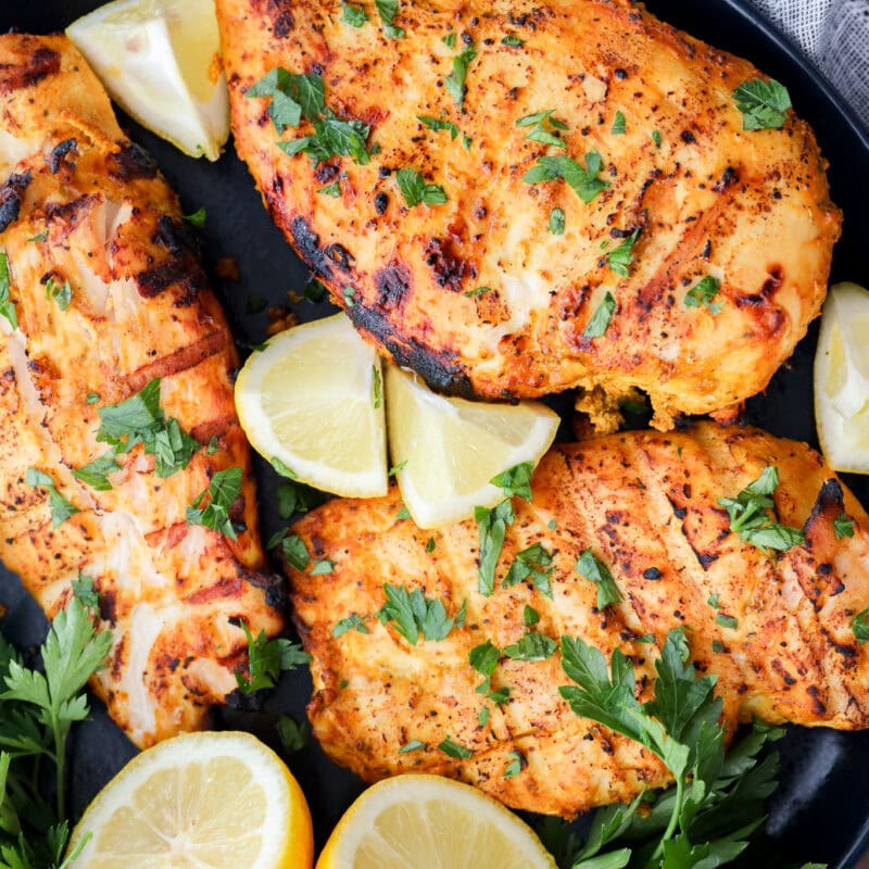 Three grilled, seasoned chicken breasts garnished with fresh parsley are arranged on a black plate with lemon wedges and parsley sprigs. A gray napkin is partially visible in the background.