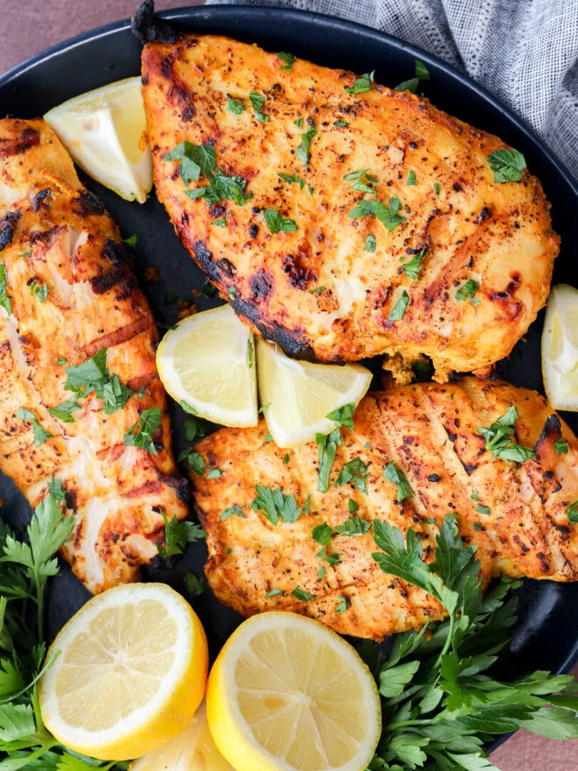 Three grilled, seasoned chicken breasts garnished with fresh parsley are arranged on a black plate with lemon wedges and parsley sprigs. A gray napkin is partially visible in the background.