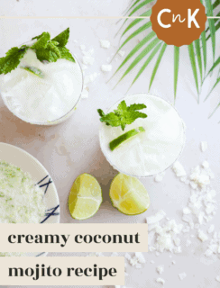 Two glasses of creamy Coconut Mojito with mint and lime garnish sit on a light surface next to fresh lime halves, coconut flakes, a palm leaf, and a bowl of coconut, with text reading creamy Coconut Mojito recipe.