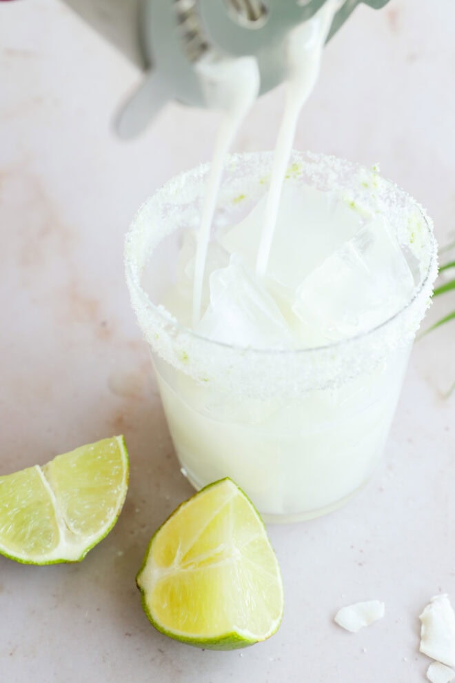A glass rimmed with salt and filled with ice cubes is being topped with a milky white Coconut Mojito. Two lime wedges are placed on the surface next to the glass.