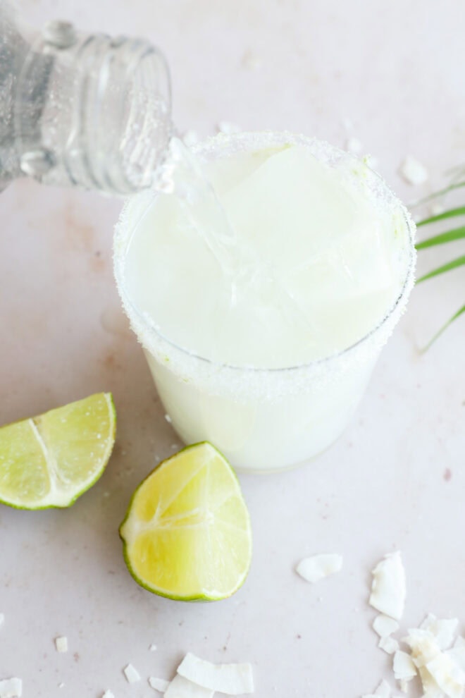 A glass with a salted rim and ice cubes is being filled with a clear club soda, surrounded by lime wedges and coconut flakes—perfect for serving a refreshing Coconut Mojito on a light surface.