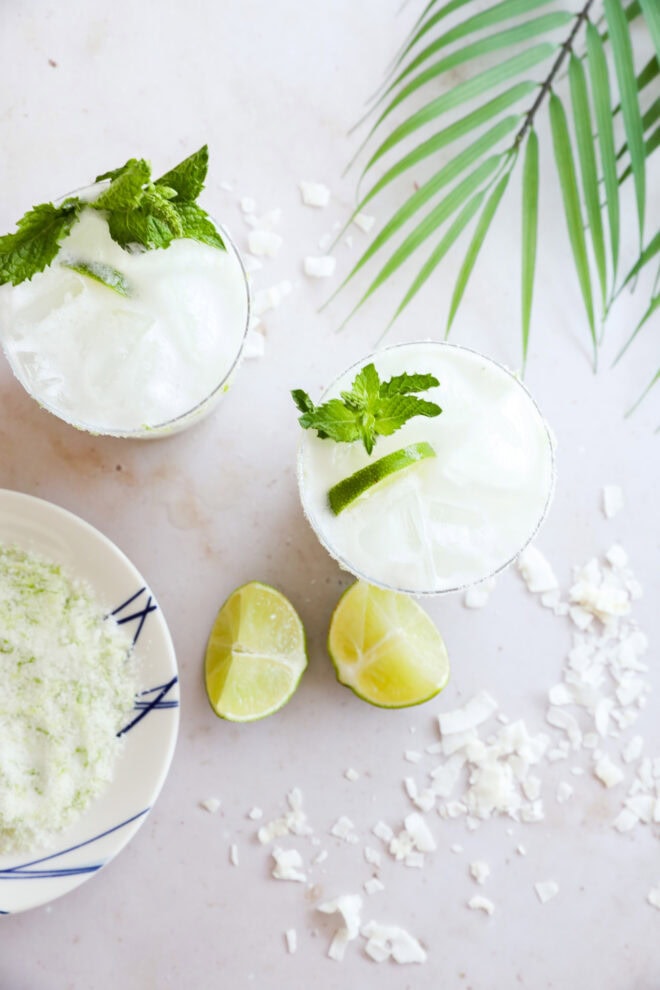 Two glasses of iced Coconut Mojito sit on a white surface with mint and lime garnish, next to lime halves, coconut flakes, a plate with a lime-salt mixture, and a palm leaf.