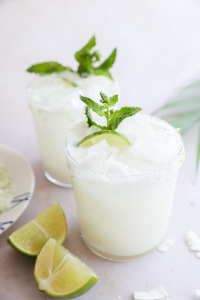 Two glasses of Coconut Mojito with ice, garnished with lime slices and mint leaves. Lime wedges and a bowl of salt sit beside the refreshing drinks on a light surface.