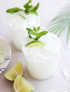 Two glasses of a white, frothy drink with ice, are garnished with lime wedges and fresh mint. Lime wedges and coconut flakes sit on the white surface nearby, with a palm leaf in the background.