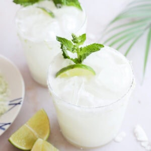 Two glasses of a white, frothy drink with ice, are garnished with lime wedges and fresh mint. Lime wedges and coconut flakes sit on the white surface nearby, with a palm leaf in the background.