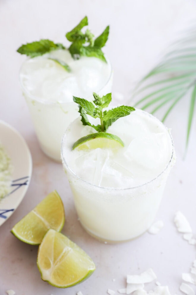 Two glasses of a white, frothy drink with ice, are garnished with lime wedges and fresh mint. Lime wedges and coconut flakes sit on the white surface nearby, with a palm leaf in the background.
