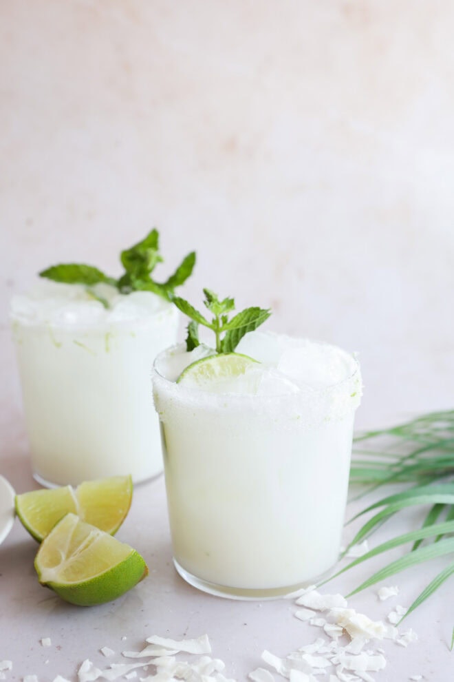 Two glasses of creamy Coconut Mojito with ice, garnished with mint leaves and lime wedges, sit on a light surface. Lime wedges and coconut flakes are scattered nearby. The background is neutral and minimalistic.