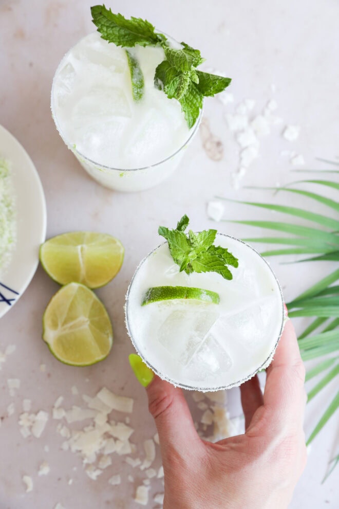 A hand holds a glass of cold, white Coconut Mojito garnished with lime slices and mint. Another similar drink, lime wedges, and coconut flakes are on a light surface nearby, along with a palm leaf decor.