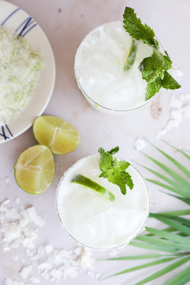 Overhead view of two glasses filled with a white, frothy cocktail garnished with mint and lime slices. Nearby are lime wedges, a plate with a powdery substance, and a green palm leaf on a light surface.