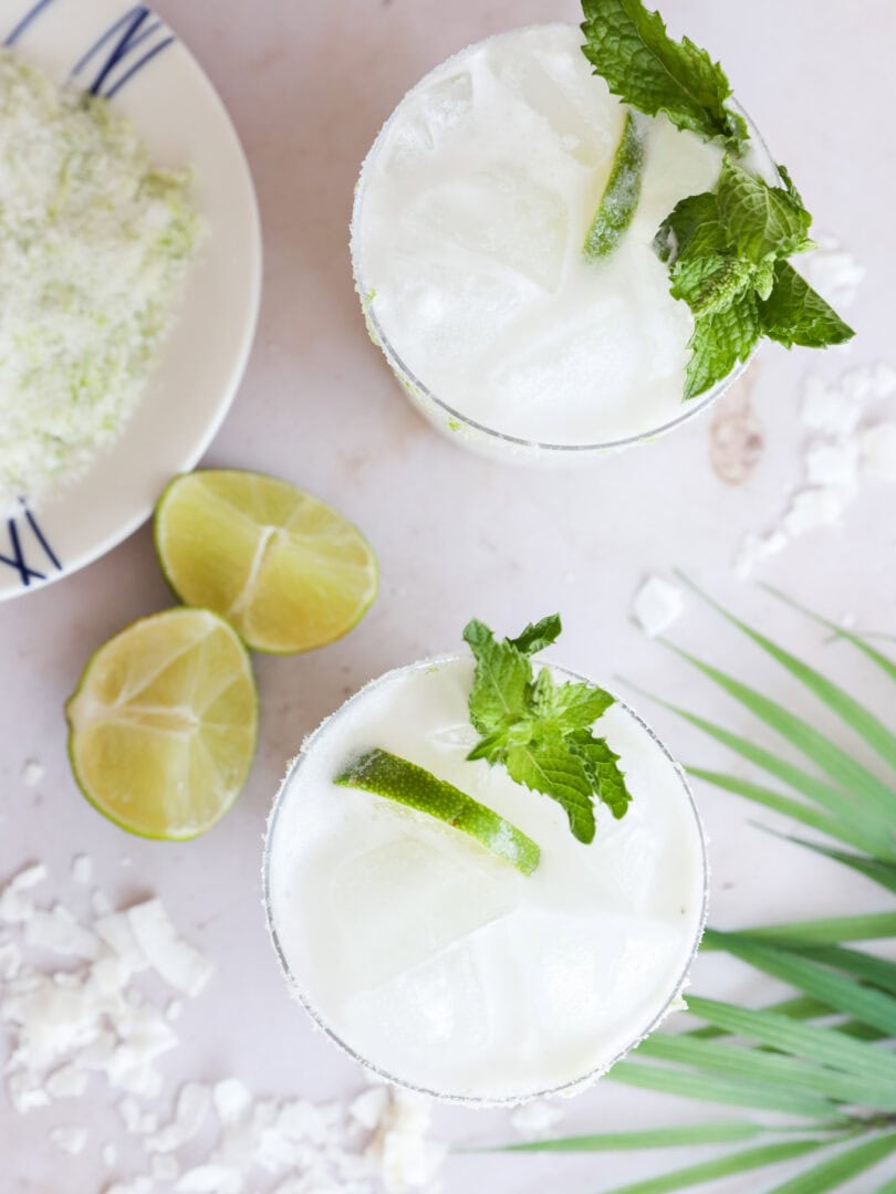 Overhead view of two glasses filled with a white, frothy cocktail garnished with mint and lime slices. Nearby are lime wedges, a plate with a powdery substance, and a green palm leaf on a light surface.