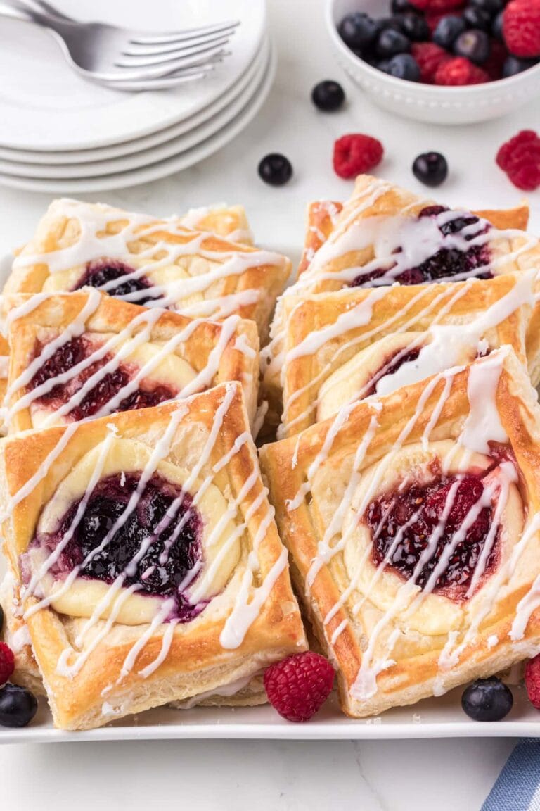 A plate of square pastries made from puff pastry dessert recipes, filled with cream cheese and raspberry jam, and drizzled with white icing. Fresh raspberries and blueberries are scattered around, with bowls of berries and stacked plates in the background.