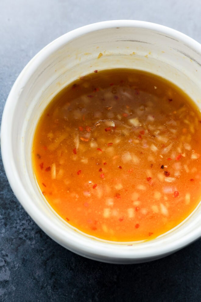 A white bowl filled with a reddish-orange sauce containing visible bits of minced garlic and red chili, set on a dark surface.