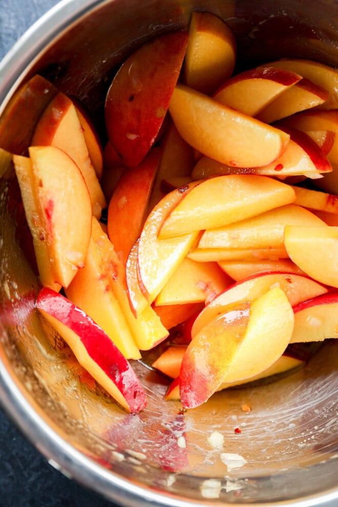 Sliced fresh peaches with red and yellow skin in a metal mixing bowl, lightly coated with a glaze or dressing.