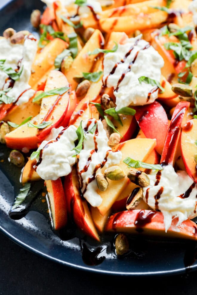 A close-up of a fresh salad with sliced peaches and nectarines, dollops of creamy cheese, chopped basil, pistachios, and drizzles of balsamic glaze, served on a dark plate.