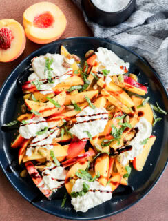 A black plate with sliced peaches, strips of red bell pepper, dollops of creamy cheese, chopped basil, pistachios, and a drizzle of balsamic glaze, next to a halved peach and a small bowl of flaky salt.