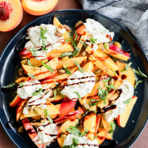 A black plate with sliced peaches, strips of red bell pepper, dollops of creamy cheese, chopped basil, pistachios, and a drizzle of balsamic glaze, next to a halved peach and a small bowl of flaky salt.