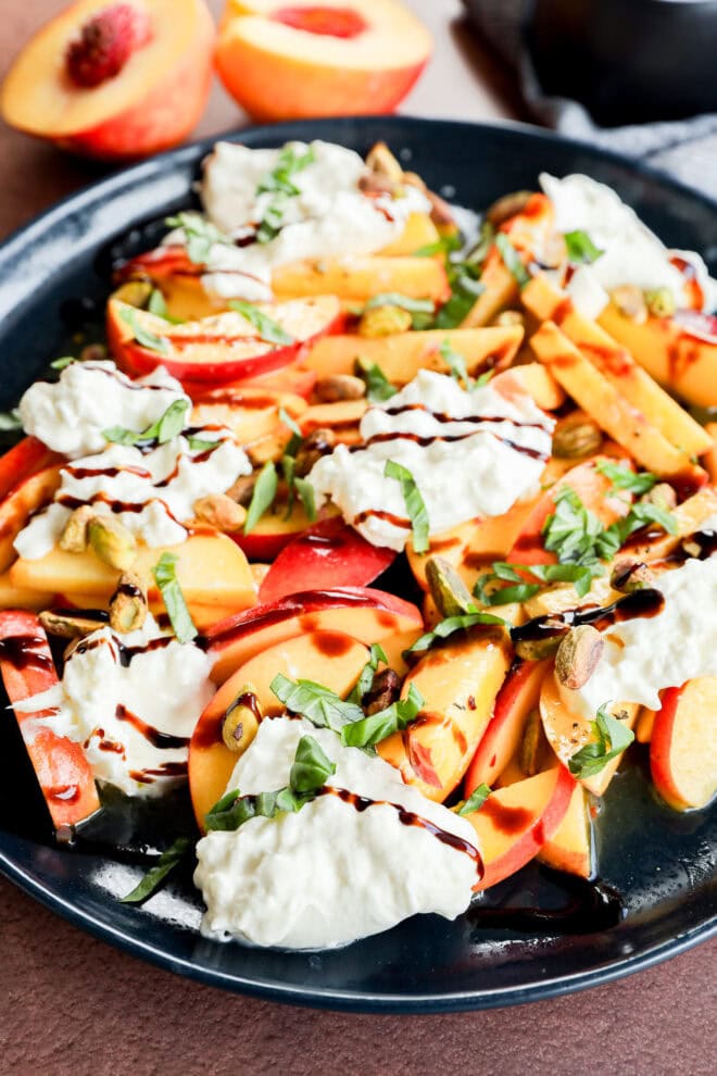 A plate of sliced peaches, fresh mozzarella or burrata, chopped pistachios, fresh basil, and a drizzle of balsamic glaze, with halved peaches in the background.