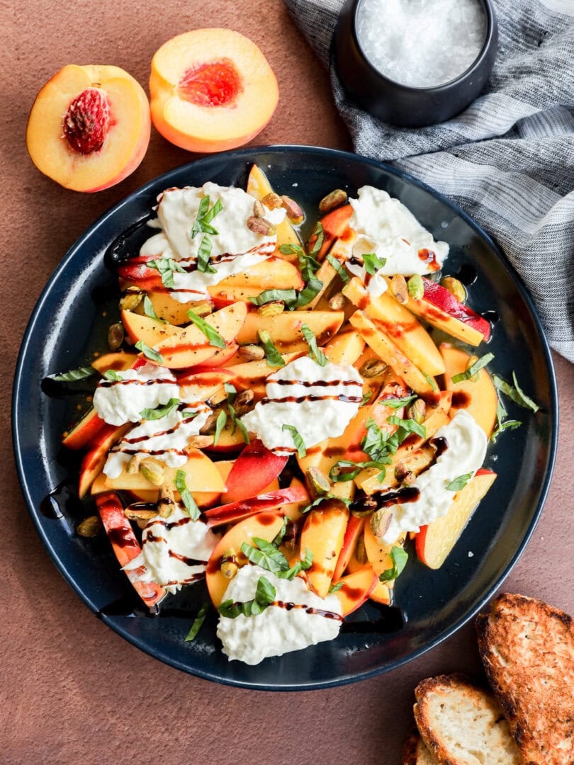 A plate of fresh salad with sliced peaches, burrata cheese, basil, and a drizzle of balsamic glaze, served with toasted bread. A halved peach and a bowl of salt are nearby on a brown surface.