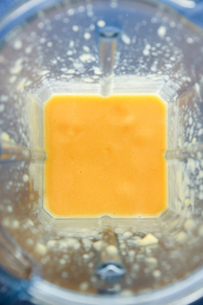 A top-down view of a blender containing a smooth, orange peach puree, with some splatters of the mixture on the sides of the blender jar.