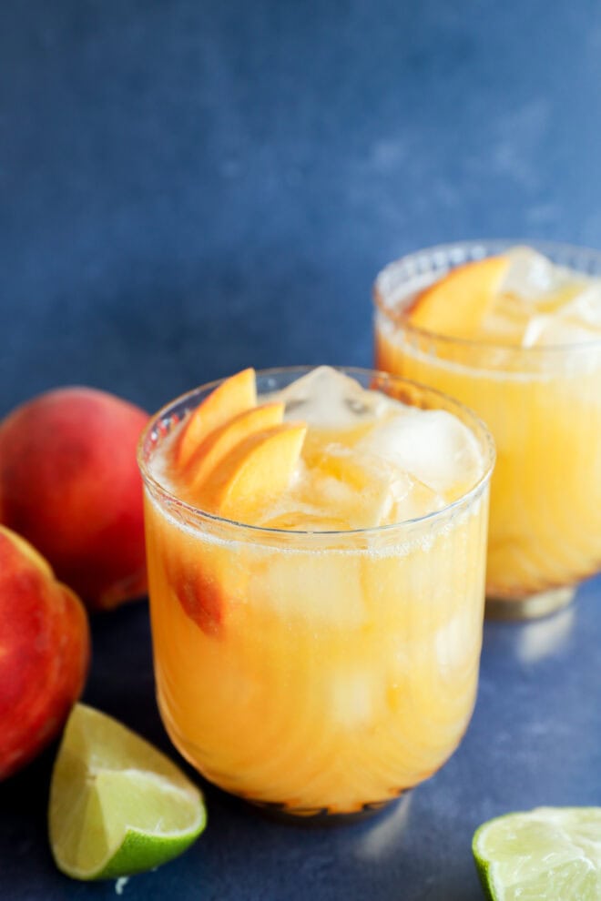 A glass of iced peach margarita garnished with fresh peach slices sits on a blue surface, with another similar glass, whole peaches, and lime wedges in the background.