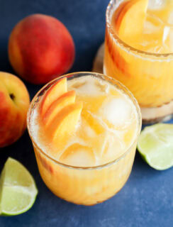 Two glasses of iced peach margarita cocktails garnished with peach slices, surrounded by fresh peaches and lime wedges on a blue surface.