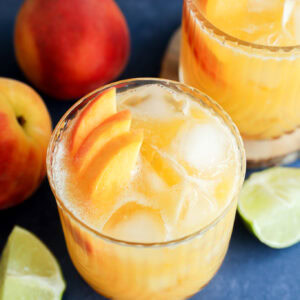 Two glasses of iced peach margarita cocktails garnished with peach slices, surrounded by fresh peaches and lime wedges on a blue surface.