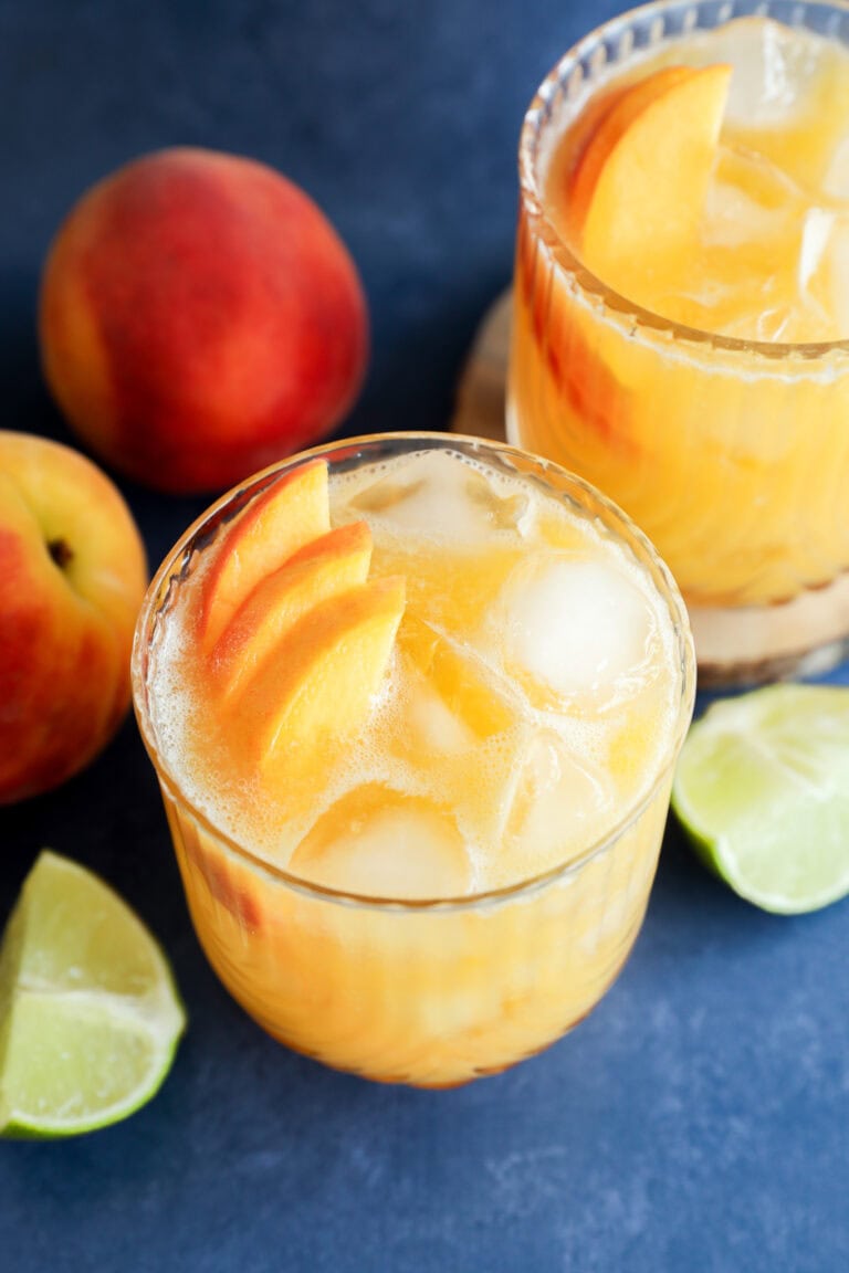 Two glasses of iced peach margarita cocktails garnished with peach slices, surrounded by fresh peaches and lime wedges on a blue surface.