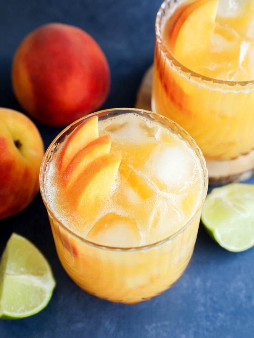 Two glasses of iced peach margarita cocktails garnished with peach slices, surrounded by fresh peaches and lime wedges on a blue surface.