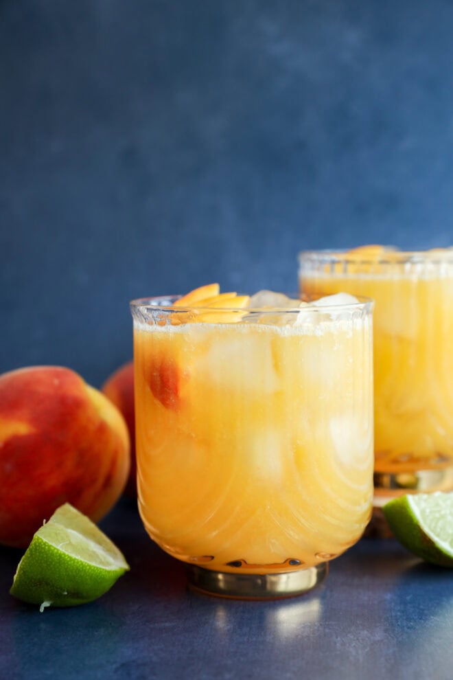 Two glasses of peach margarita, garnished with peach slices, sit on a dark surface. Fresh peaches and a halved lime are nearby, set against a blurry blue background.