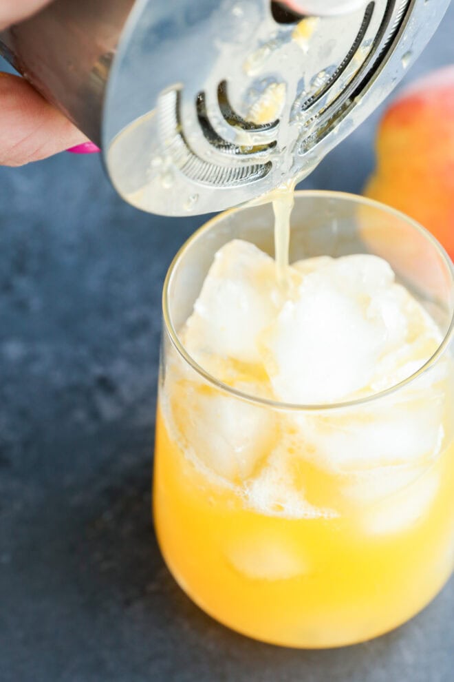 A close-up of a hand pouring a yellow-orange drink over ice cubes in a glass using a cocktail shaker and strainer; a peach is visible in the background on a dark surface.
