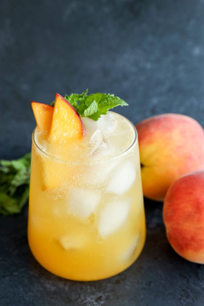 A glass filled with an iced peach-colored drink, garnished with peach slices and mint, sits on a dark surface next to two whole peaches and some fresh mint leaves.