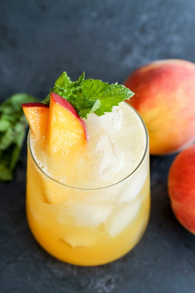 A glass of peach-colored iced drink garnished with peach slices and a sprig of mint, with fresh peaches and mint leaves in the background on a dark surface.