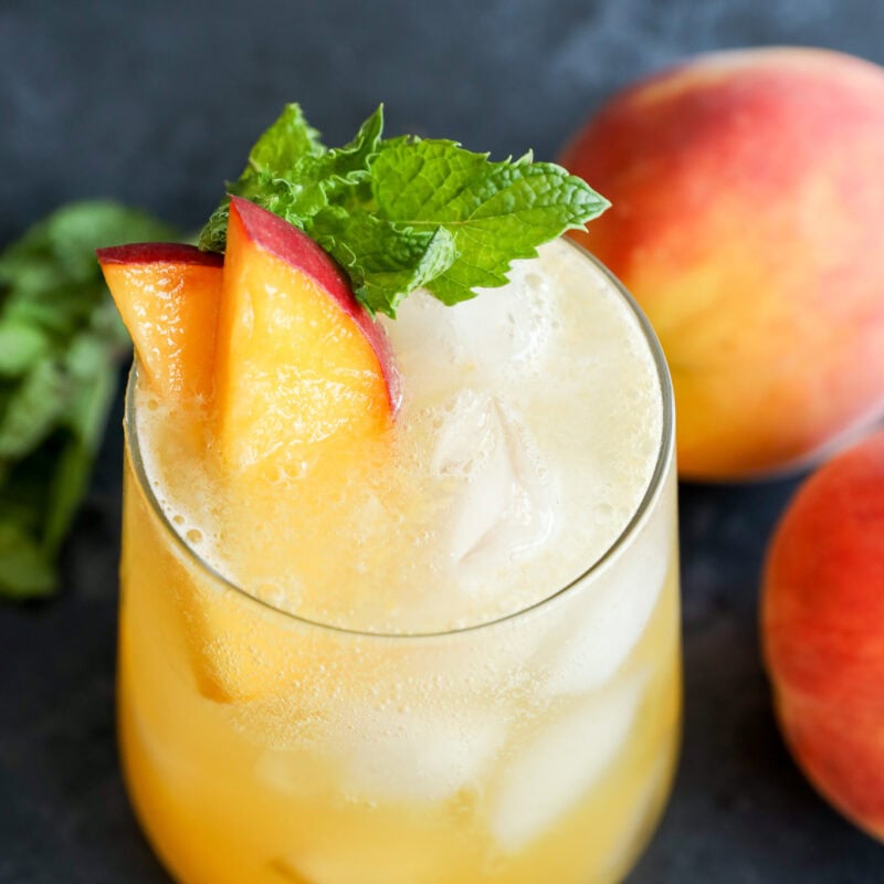 A glass of peach-colored iced drink garnished with peach slices and a sprig of mint, with fresh peaches and mint leaves in the background on a dark surface.