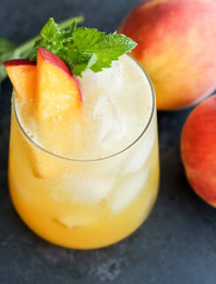 A glass of iced peach drink garnished with peach slices and mint leaves, placed next to two whole peaches and a small bunch of mint on a dark surface.