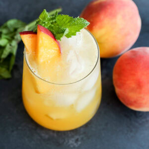 A glass of iced peach drink garnished with peach slices and mint leaves, placed next to two whole peaches and a small bunch of mint on a dark surface.