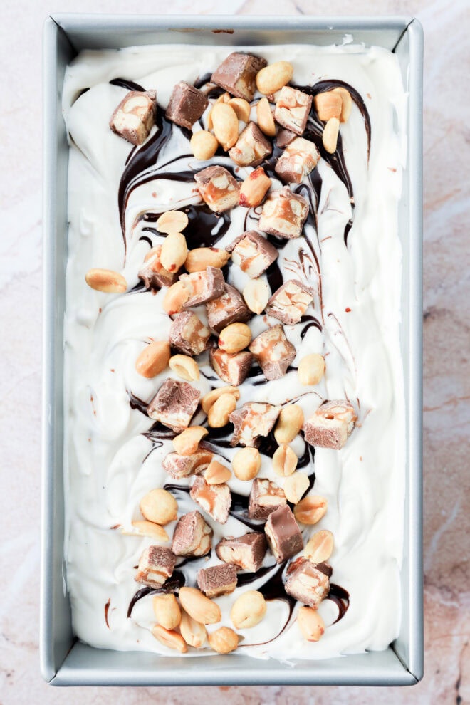 A rectangular pan filled with swirled vanilla dessert topped with chopped chocolate candy bars, peanuts, and ribbons of chocolate syrup.