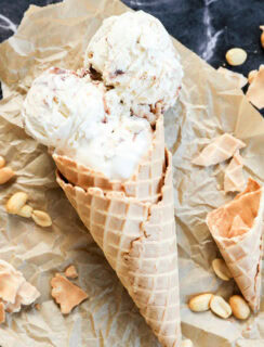 Two scoops of Snickers Ice Cream in a waffle cone rest on crumpled parchment paper, surrounded by scattered peanuts and broken pieces of cone on a dark marble surface.