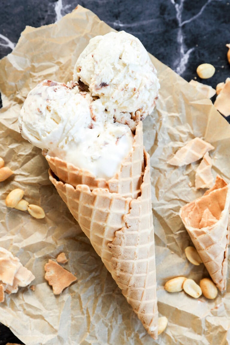 Two scoops of Snickers Ice Cream in a waffle cone rest on crumpled parchment paper, surrounded by scattered peanuts and broken pieces of cone on a dark marble surface.