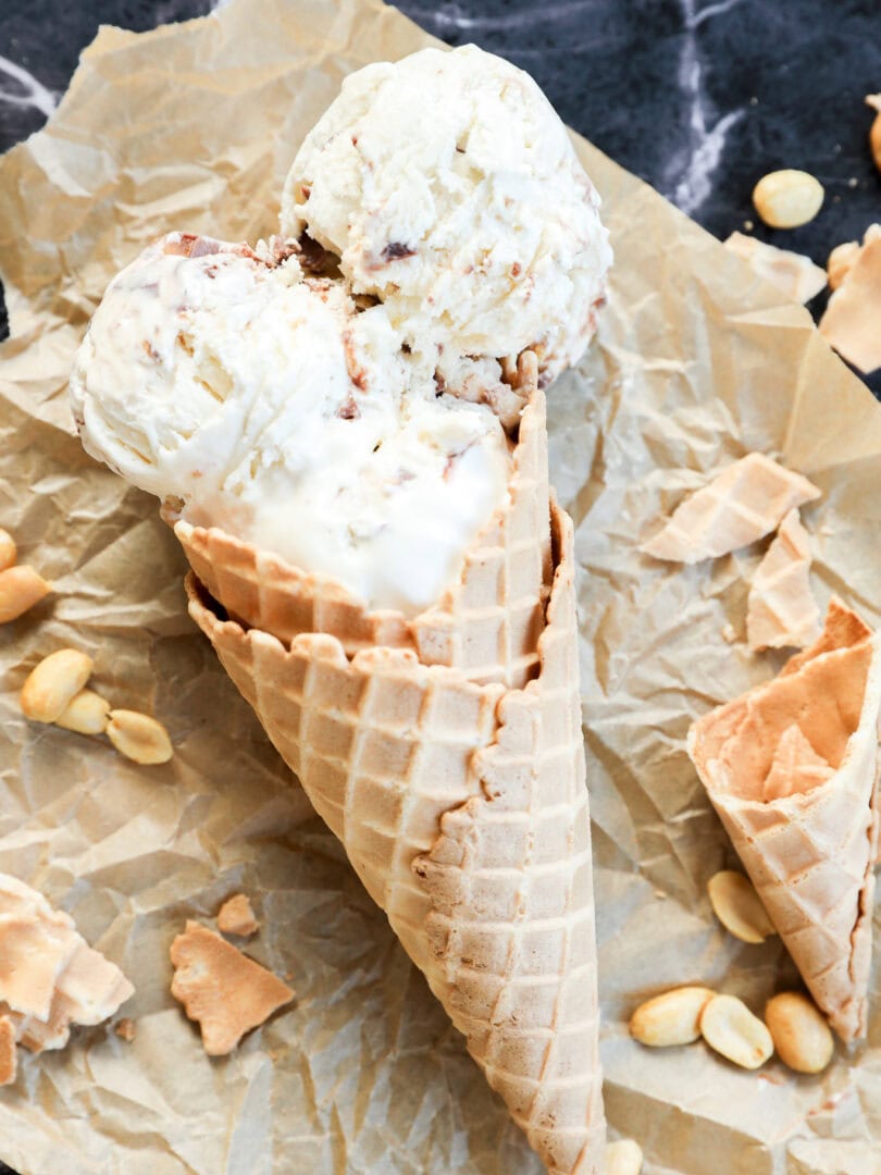 Two scoops of Snickers Ice Cream in a waffle cone rest on crumpled parchment paper, surrounded by scattered peanuts and broken pieces of cone on a dark marble surface.