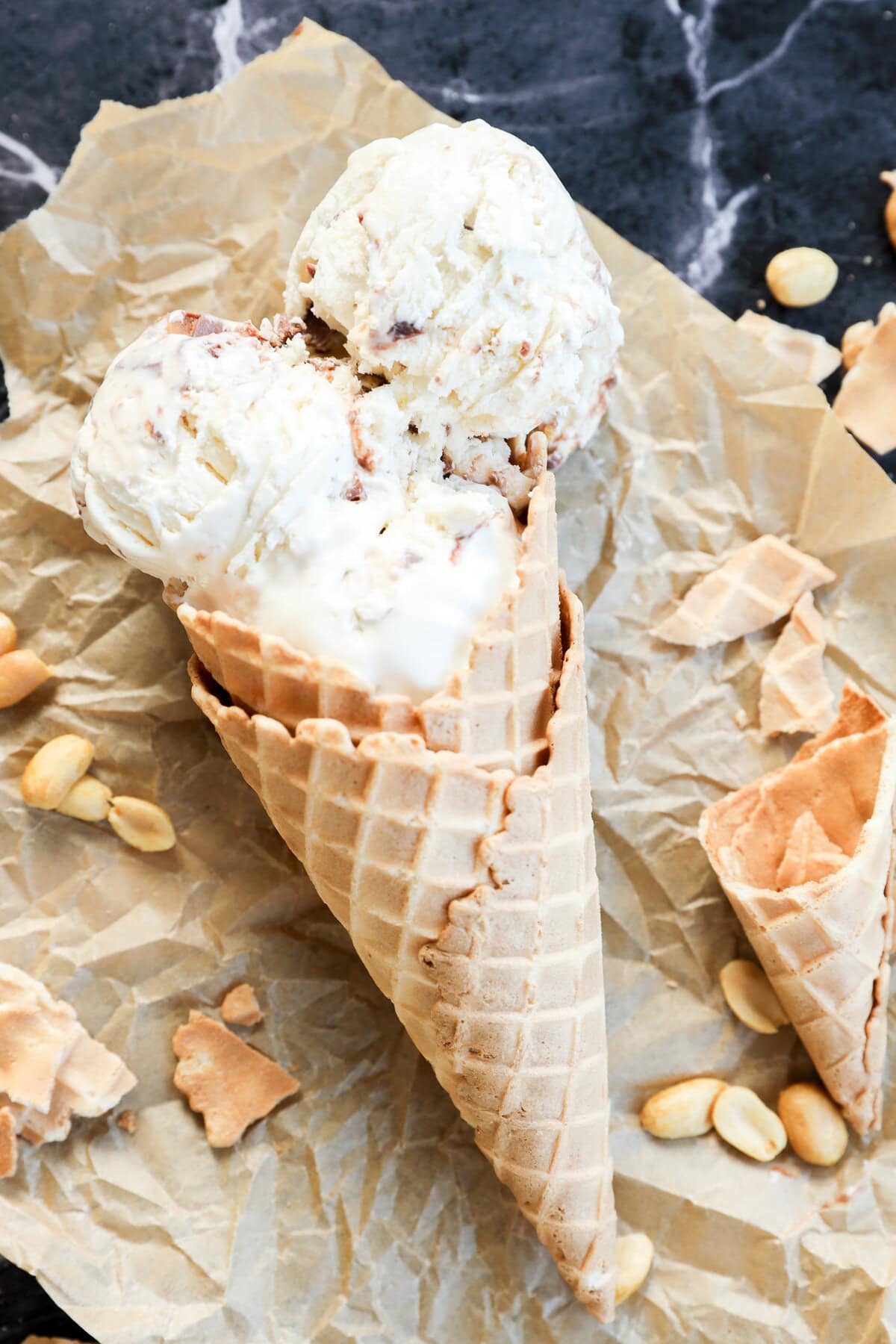 Two scoops of Snickers Ice Cream in a waffle cone rest on crumpled parchment paper, surrounded by scattered peanuts and broken pieces of cone on a dark marble surface.